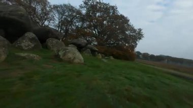 Bir sonbahar günü bulutlu bir gökyüzü altında yeşil bir manzaranın ortasında antik megalitik mezar inşaatını ortaya çıkaran neolitik dolmenlerin etrafında uçan etkileyici Fpv drone görüntüleri.