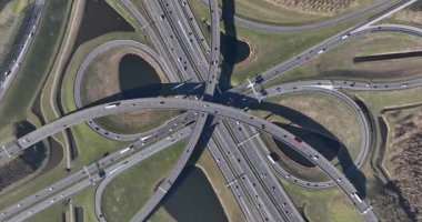 Yukarıdan aşağıya yüksek açılı, havadan çekim. Karmaşık bir otoyol kavşağında yavaşça dönüyor. Birden fazla üst geçit ve rampa var.