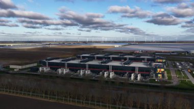 Geniş bir güneş çiftliği ve rüzgar türbinleri dahil sürdürülebilir enerji kaynaklarına sahip büyük bir veri merkezinin hava görüntüsü parçalı bulutlu bir gökyüzü altında temiz elektrik üretiyor.