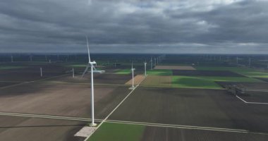Uçsuz bucaksız bir rüzgar tarlasının havadan görünüşü. Dönen türbinler, sürdürülebilir ve temiz enerji üretiyor. Yayılan tarım alanları ve kırsal alanlarda dramatik bulutlu bir gökyüzüne karşı kuruluyor.