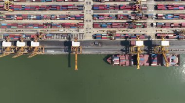 Yoğun bir endüstriyel limandan kalkan konteynırlarla dolu büyük bir kargo gemisinin yukarıdan aşağıya bakan görüntüsü.