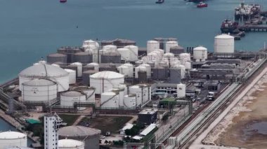 Büyük beyaz depolama tankları, karmaşık boru hattı altyapısı ve etrafındaki mavi denizde yakıt nakli için bekleyen kargo gemileri olan büyük bir sahil petrol rafinerisinin havadan görüntüsü.