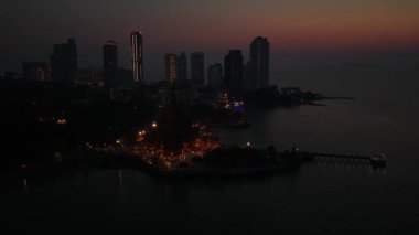 Güzel bir alacakaranlıkta, kıyı boyunca aydınlık gökdelenleri olan modern bir şehrin gökyüzü perspektifi, sakin denizi ve renkli gökyüzü altında küçük bir iskeleyi gözler önüne seriyor.