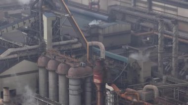 Paslı fırınları ve dumanlı bacaları olan büyük, yaşlanan bir çelik fabrikasının havadan görünüşü yoğun beyaz dumanlar salıyor, engin endüstriyel bir komplekste ağır hava kirliliğine yol açıyor.