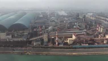 Nehrin yanında bulunan çok sayıda bina, baca bacası ve boru hatlarının bulunduğu büyük bir endüstriyel kompleksin hava görüntüsü, üretim sırasında sisli atmosfere duman ve buhar yayıyor.