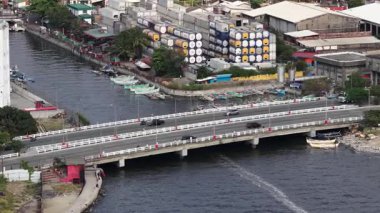 Kalabalık bir kentsel alanda beton bir köprüden geçen araçların yüksek açılı görüntüsü, bankalar boyunca demirlemiş geleneksel tekneler ve arka planda istiflenmiş kargo konteynırları ile