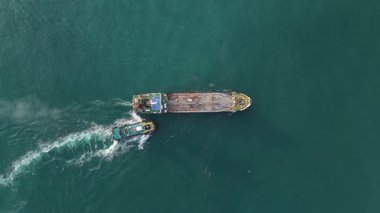 Güçlü bir römorkör, deniz lojistiği, ulaşım ve endüstriyel nakliye operasyonlarını gösteren, sakin, yeşil okyanus boyunca boş bir mavnayı hareket ettiren yüksek açılı hava görüntüsü.