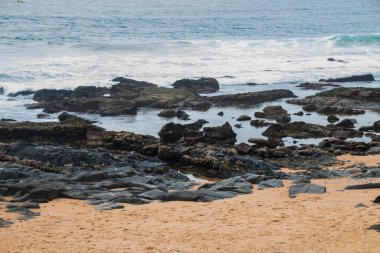 Kıyı şeridinde siyah kayalar ve temiz kumsal kumları.