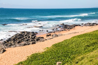 Sörf ve siyah kayalarla kaplı bir kum tepesi.