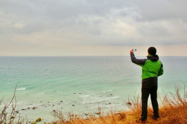 Yeşil su geçirmez ceketli adamı coastliny fotoğrafını tarafından akıllı telefon alır. Soğuk rüzgarlı gün denizde.