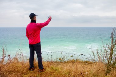 Beyzbol kap ve pembe windcheater turist fotoğraf cep telefonu alır. Okyanus, rüzgarlı gün