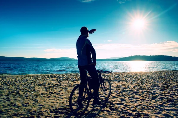 年轻男子在海滩上的日落背景和骑自行车的人轮廓在蔚蓝的天空。在湖的赛季结束. — 图库照片