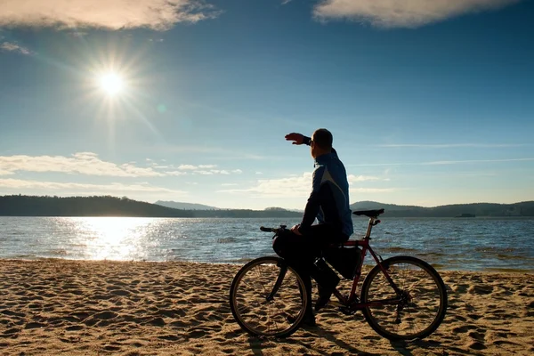 Młody człowiek rowerzysta siedzieć na rower, niebieskie tło niebo i zachód słońca na plaży. Koniec sezonu nad jeziorem — Zdjęcie stockowe