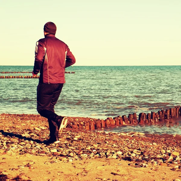 Tall man in dark sportswer running and exercising on stony beach at breakwater.