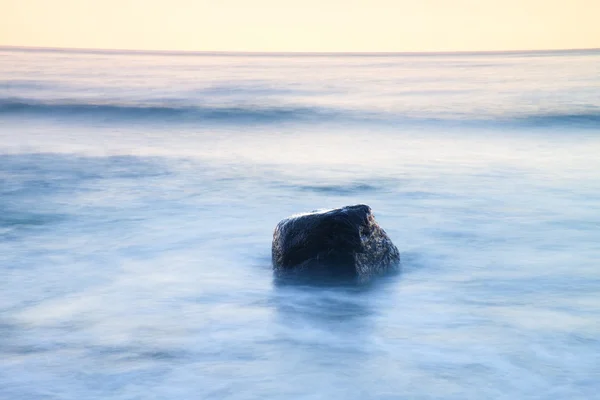 海の静かな朝でロマンチックな雰囲気。大きな岩が滑らかな波状の海から突き出ています。ピンクの地平線 — ストック写真