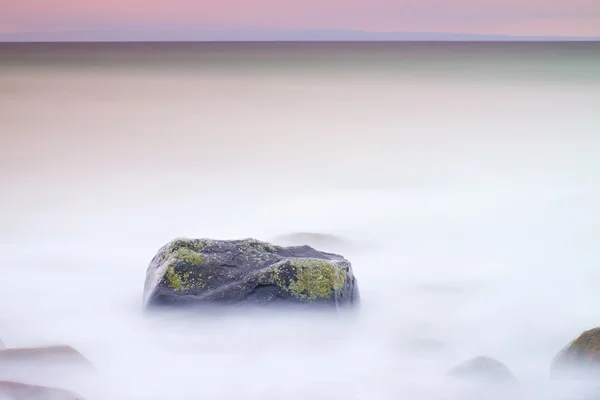 海の静かな朝でロマンチックな雰囲気。大きな岩が滑らかな波状の海から突き出ています。ピンクの地平線 — ストック写真