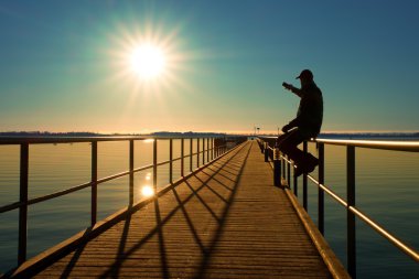 Adam iskele üzerinde oturup sabah denize tadını çıkarın. Güneşli açık mavi gökyüzü, yumuşak su seviyesi