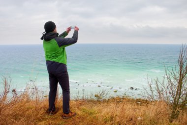 Yeşil su geçirmez ceketli adamı coastliny fotoğrafını tarafından akıllı telefon alır. Soğuk rüzgarlı gün denizde.