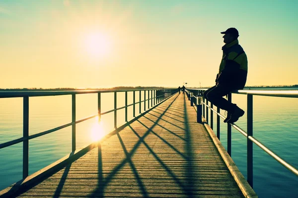 橋脚手すり工事で暖かいジャケットと野球キャップ座って男し、海で朝をお楽しみください。. — ストック写真