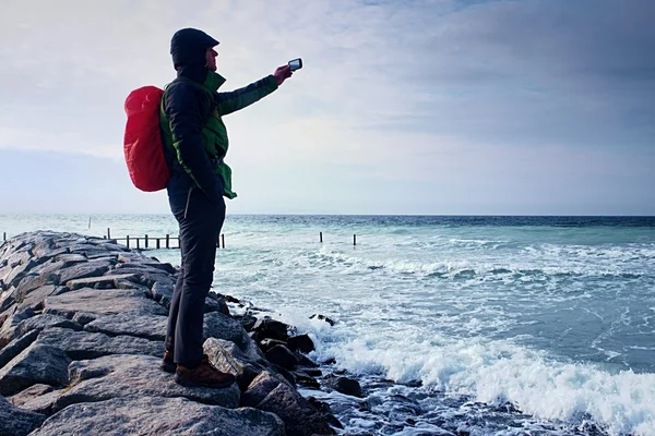 Caminante en el muelle fotografía mar mañana. Turista con teléfono inteligente en la mano, mochila en impermeable rojo. Fantástica mañana — Foto de Stock