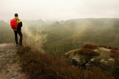 Tourist with backpack takes photos with smart phone of rainy vally. Dreamy foggy valley below clipart