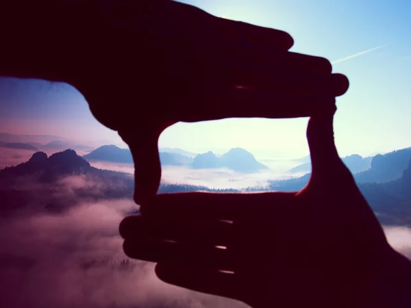 Nahaufnahme von Händen, die eine Rahmengeste machen. blauer Nebel unter felsigen Gipfeln. Sonnenaufgang im Frühling in den Bergen. — Stockfoto