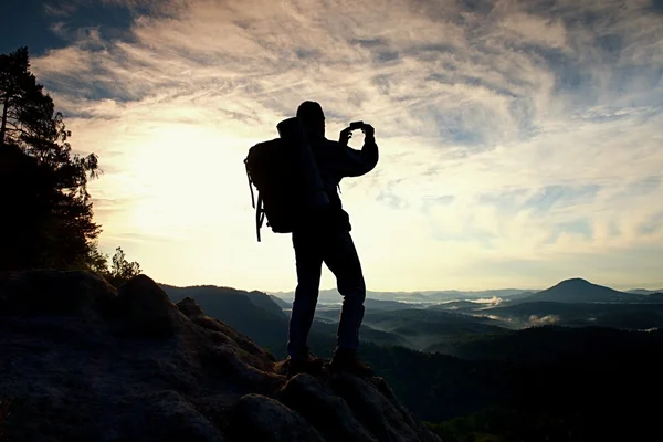 Turista con zaino pesante scatta foto con smartphone. L'uomo sulla cima rocciosa. Valle nebbiosa da sogno sotto — Foto Stock