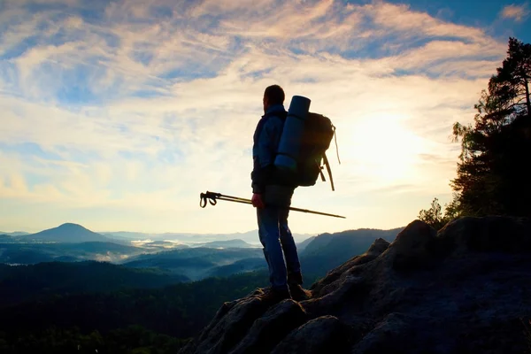 Туристический гид с шестом в руке. Турист со спортивным рюкзаком стоит на скалистой точке обзора над туманной долиной. Солнечный весенний рассвет — стоковое фото
