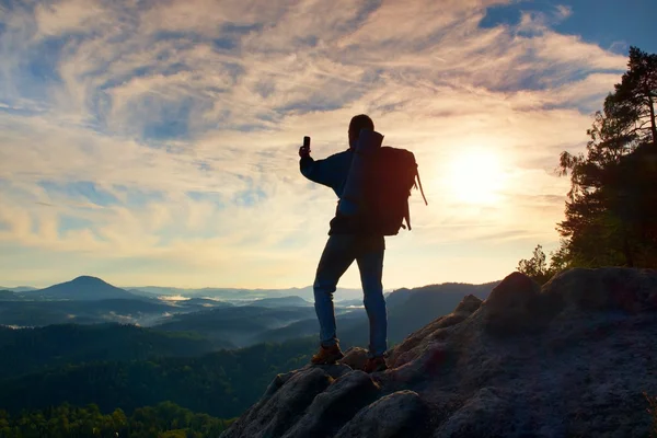 Турист с тяжелым рюкзаком фотографируется со смартфоном. Человек на скалистой вершине. Мечтательная туманная долина внизу — стоковое фото