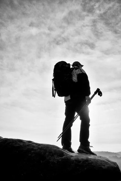 Silueta turista s tyčí v ruce. Tramp s velkým batohem stojí na skalní vyhlídka nad mlhavé údolí. Jarní Slunečné svítání — Stock fotografie