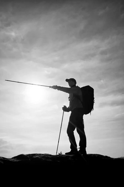 Guia turístico mostrar o caminho certo com pólo na mão. Caminhante com mochila desportiva stand no ponto de vista rochoso acima do vale enevoado . — Fotografia de Stock