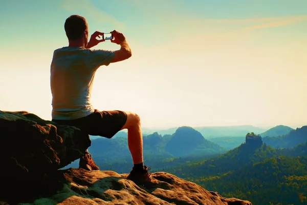 L'escursionista seduto sta scattando foto con lo smartphone sulla scogliera tagliente della montagna all'alba . — Foto Stock