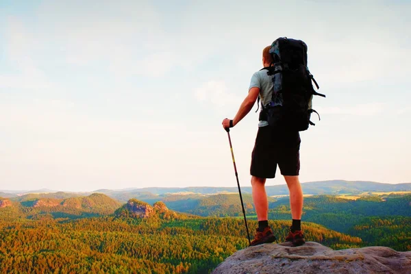 Mochileiro alto com postes na mão. Noite de verão ensolarada em montanhas rochosas. Caminhante com grande mochila no ponto de vista rochoso acima do vale . — Fotografia de Stock