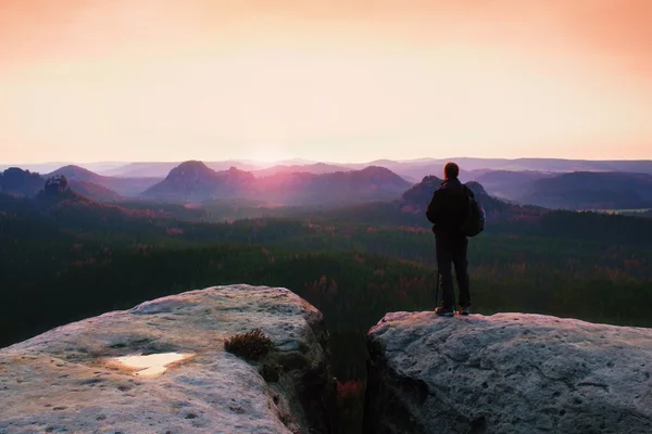 Guía turística sobre roca con poste en mano. Caminante con mochila deportiva de pie en el acantilado sobre el valle brumoso. Soleado amanecer de primavera —  Fotos de Stock