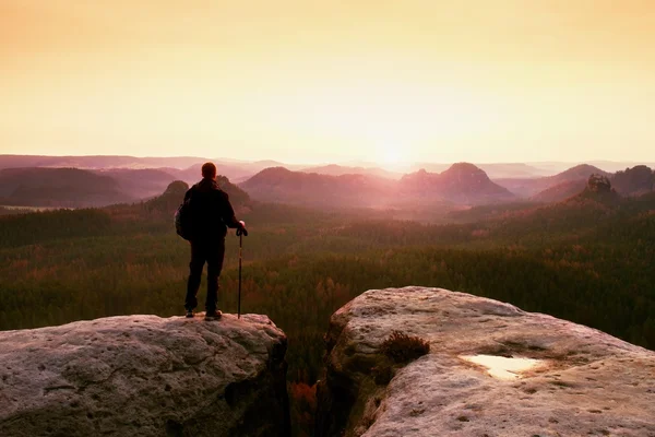 Turista com mochila e postes no pico rochoso olhando para o sol. Vale nebuloso sonhador abaixo — Fotografia de Stock