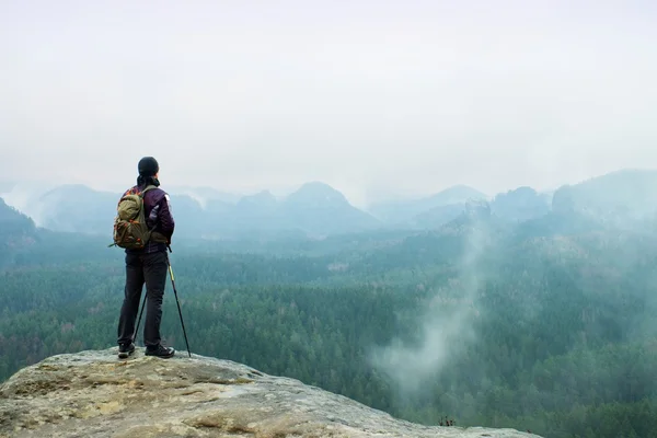 Caminante con mochila y postes en roca del acantilado que mira sobre el valle brumoso y nebuloso de la primavera a Horizonte . —  Fotos de Stock