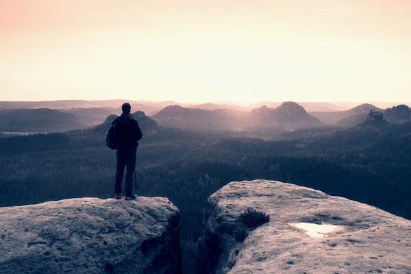 Tourist with backpack and poles on rocky peak looking into sun. Dreamy fogy valley below — Stock Photo, Image