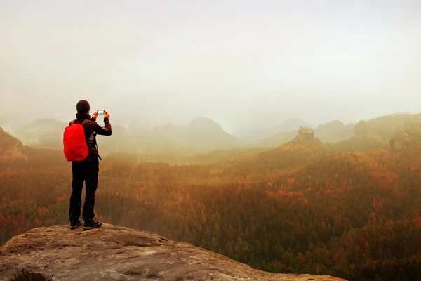 Tourist with backpack takes photos with smart phone of rainy vally. Dreamy foggy valley below — Stock Photo, Image