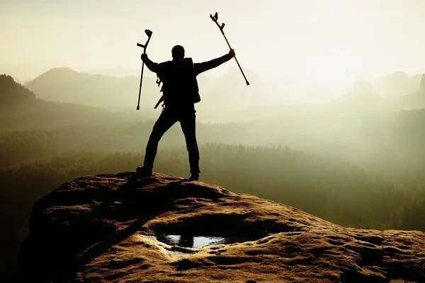 Hiker with broken leg in immobilizer and medicine pole above head achieved rocky summit. Deep misty valley bellow — Stock Photo, Image