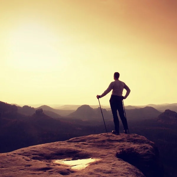 Ginger hair man in grey t-shirt and dark trekking trousers on sharp rock. Tourist with pole above misty valley. — Stock Photo, Image