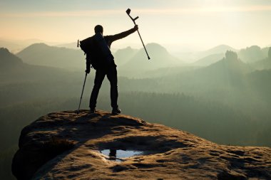 Tourist with  medicine crutch above head achieved mountain peak. Hiker with broken leg in immobilizer.  Deep misty valley bellow silhouette of man with hand in air. Spring daybreak clipart