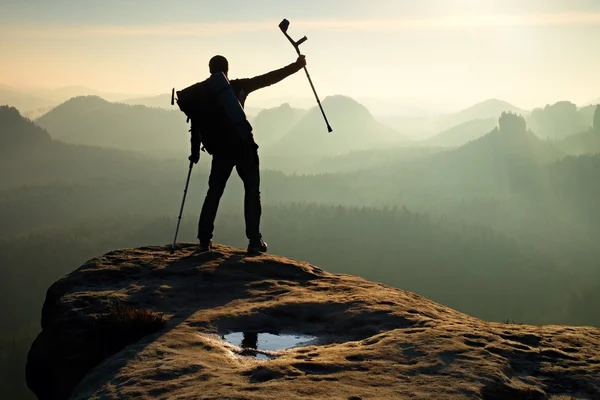 Tourist with  medicine crutch above head achieved mountain peak. Hiker with broken leg in immobilizer.  Deep misty valley bellow silhouette of man with hand in air. Spring daybreak — Stock Photo, Image