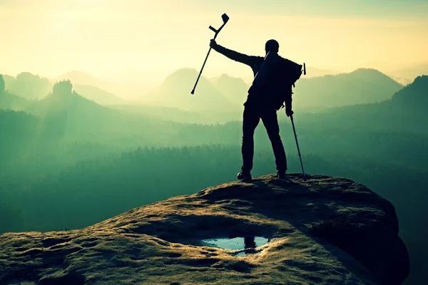 Turista con medicina muleta por encima de la cabeza alcanzado pico de montaña. Caminante con la pierna rota en inmovilizador. Profundo valle brumoso silueta de hombre con la mano en el aire. Amanecer de primavera — Foto de Stock