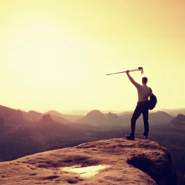 Tourist with backpack and poles on rocky peak looking into sun. Sunny daybreak in mountains, dreamy fogy valley below. — Stock Photo, Image