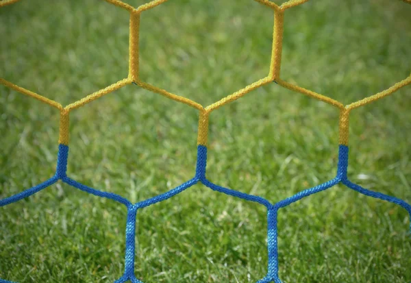 Detail of yellow blue crossed soccer nets, soccer football in goal net with grass on playground in background. Honeycomb shape. — Stock Photo, Image