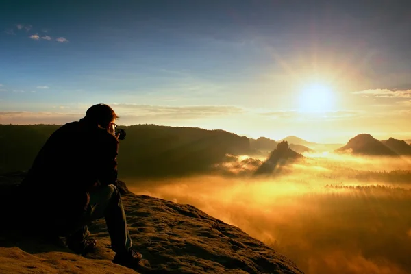 Happy photo enthusiast is  enjoying fantastic miracle of nature on cliff on rock. Dreamy fogy landscape bellow. — Stock Photo, Image