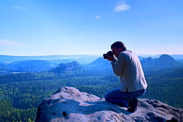 Homem de cabelo escuro está tirando foto por câmera grande espelho no pescoço no pico da montanha ao nascer do sol . — Fotografia de Stock