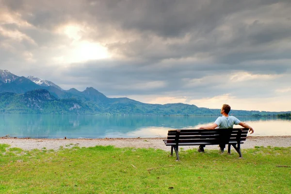 Sám muž sedí na lavici vedle azurové horské jezero. Člověk relaxovat — Stock fotografie