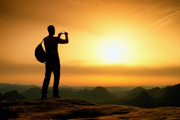 Tourist with backpack takes photos with smartphone on peak. Orange daybreak — Stock Photo, Image