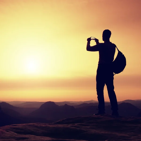 Tourist with backpack takes photos with smartphone on peak. Orange daybreak — Stock Photo, Image